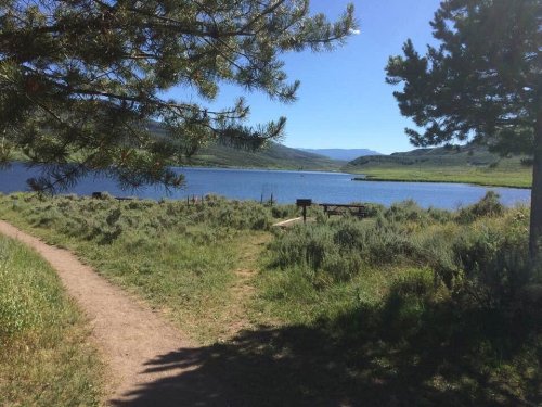 A serene lake surrounded by greenery and mountains under a clear blue sky, with a path leading to the water.