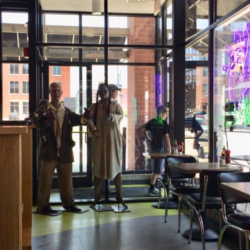 Two life-sized mannequins dressed in quirky outfits stand inside a restaurant, with a view of the street outside.
