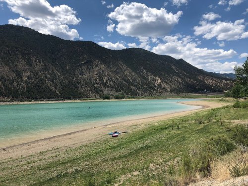 A serene lake surrounded by mountains, with a sandy shore and a few kayaks resting on the grass under a blue sky.