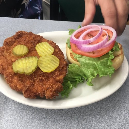 A plate with a breaded chicken patty topped with pickles, alongside a sandwich bun with lettuce, tomato, and onion slices.