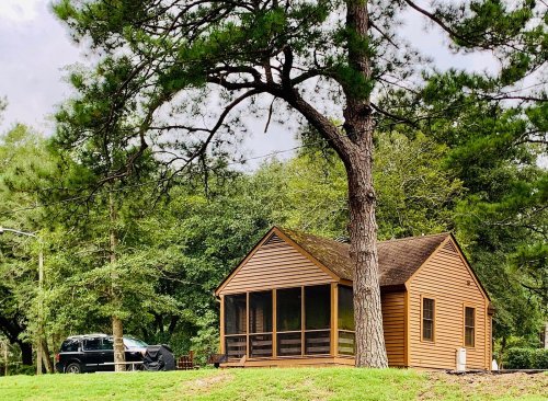 A small brown cabin with a screened porch, surrounded by trees and grass, with a black SUV parked nearby.