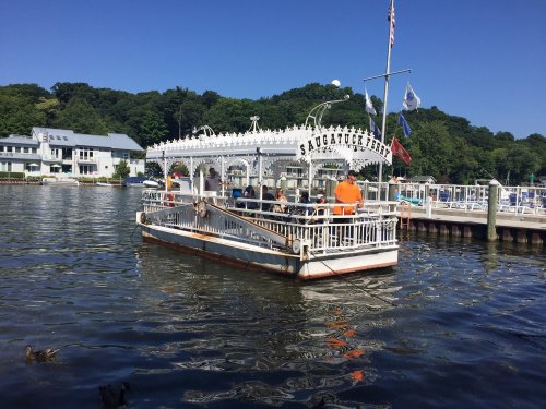 A white boat with passengers floats on a calm lake, surrounded by green trees and a dock with flags.