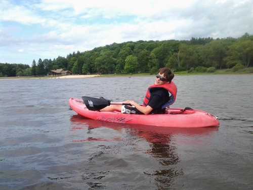 A person in a red kayak smiles while paddling on a calm lake surrounded by trees and a sandy beach in the background.