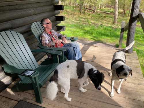 A man relaxes on a porch with two dogs, surrounded by greenery and a log cabin in the background.