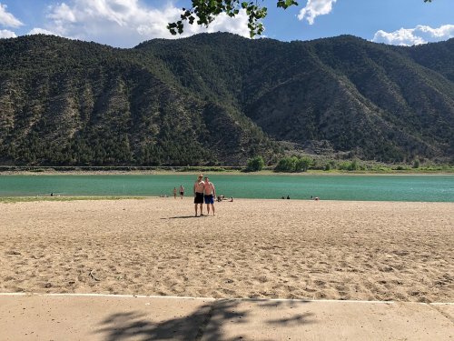 Two people stand on a sandy beach by a lake, surrounded by mountains and a clear blue sky.