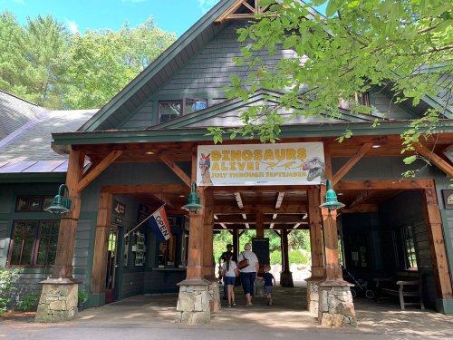 Entrance to a building with a "Dinosaurs Alive!" banner, surrounded by trees and visitors.