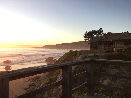 Sunset over a beach with gentle waves, a wooden railing in the foreground, and a cozy house on the shore.