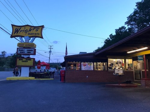 A retro diner with a bright sign, outdoor seating, and a drive-up window, set against a twilight sky.