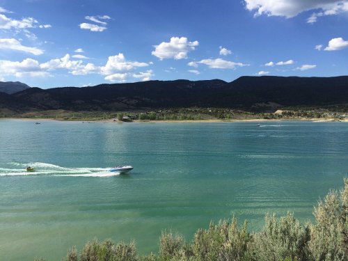 A scenic view of a calm lake with a boat cruising, surrounded by mountains and a blue sky with fluffy clouds.