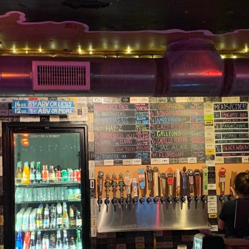 A bar menu featuring various beers, ciders, and soft drinks on a chalkboard, with taps visible below.