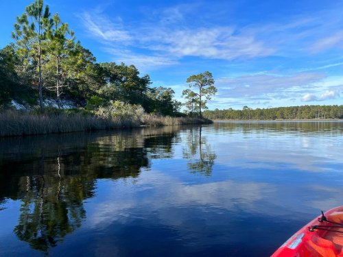 A serene waterway surrounded by trees, reflecting the sky and greenery, with a kayak in the foreground.