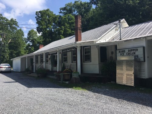 A quaint ice cream shop with a white exterior, surrounded by trees and gravel, featuring a sign for hand-dipped ice cream.