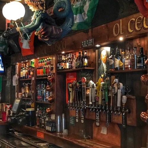 A cozy bar with a wooden counter, shelves filled with various bottles, and taps for draft beer, decorated with flags.