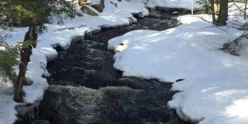 A snowy landscape with a flowing stream, surrounded by trees and patches of snow.