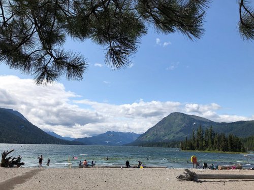 A scenic lake view with people swimming and relaxing on the beach, surrounded by mountains and trees under a blue sky.