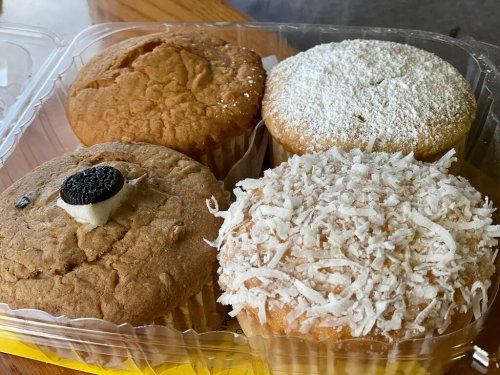 Four assorted muffins in a clear container: one with an Oreo, one with powdered sugar, and two topped with coconut.