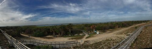 Panoramic view of a coastal landscape with wooden walkways, lush greenery, and a cloudy sky.
