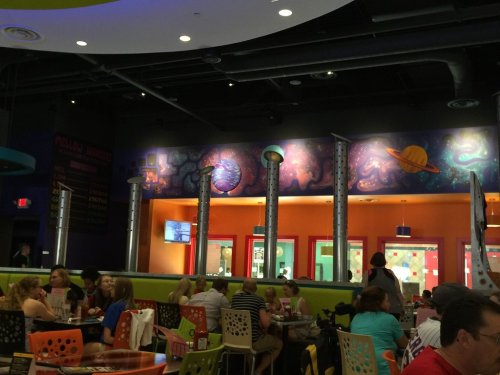 A colorful restaurant interior with a space-themed mural, featuring planets and stars, and diners enjoying their meals.