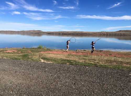 Two children play by a calm lake, one with a hoop and the other with a stick, under a clear blue sky.