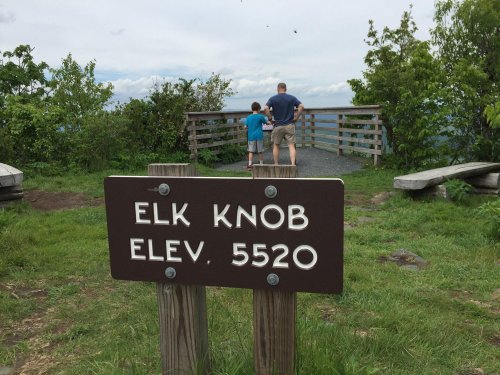 A sign reading "Elk Knob Elev. 5520" with two people walking towards a viewpoint surrounded by greenery.