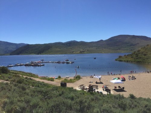 A sunny lakeside scene with people on the beach, boats on the water, and mountains in the background.