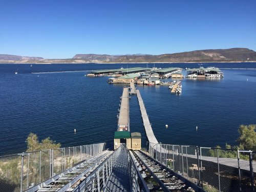 A view of a marina with docks on a calm lake, surrounded by mountains under a clear blue sky.