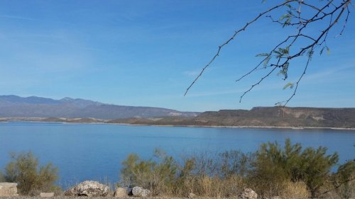 A serene lake surrounded by mountains under a clear blue sky, with sparse vegetation in the foreground.
