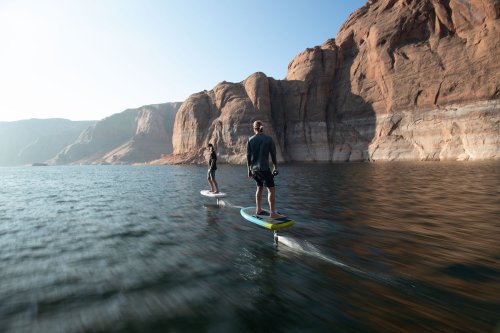 Two people surfing on hydrofoils in calm water, with rocky cliffs in the background under a clear sky.