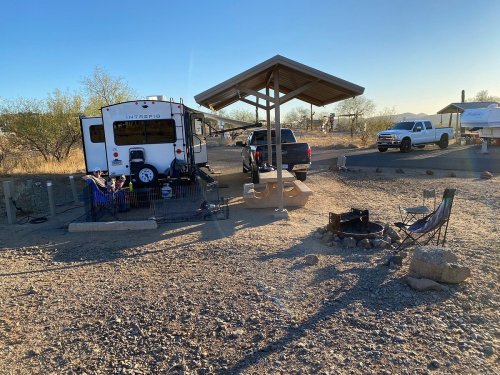 A campsite with an RV, a shaded area, a fire pit, and chairs, surrounded by desert landscape and clear blue skies.