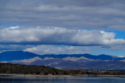 A scenic view of mountains under a cloudy sky, reflecting on a calm body of water in the foreground.