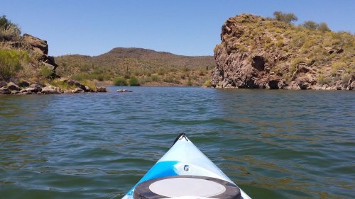 A kayak view of calm water surrounded by rocky hills and desert vegetation under a clear blue sky.
