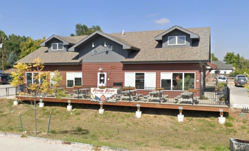 A brown wooden building with a porch, featuring a sign for "Happy Hour" and surrounded by trees and parked cars.