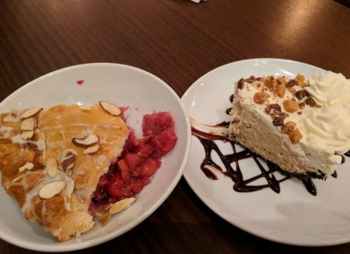A slice of cherry pie with almonds and a slice of creamy dessert topped with whipped cream and chocolate drizzle.