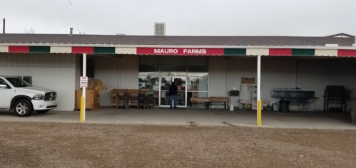 A person stands at the entrance of Mauro Farms, a building with a red and white awning, surrounded by gravel.