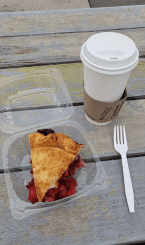 A slice of pie in a clear container next to a coffee cup on a wooden table. A plastic fork is beside the pie.