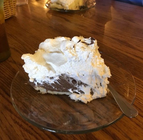 A slice of chocolate cream pie topped with whipped cream on a glass plate, resting on a wooden table.
