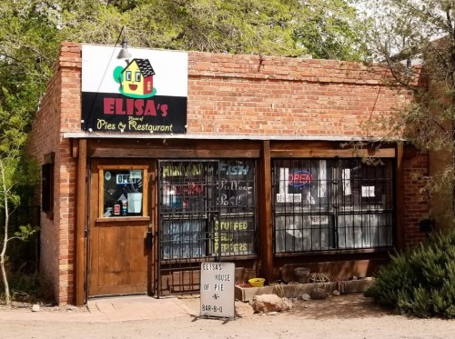 A rustic brick building with a sign reading "Elisa's Pies & Restaurant," featuring an open door and colorful window displays.