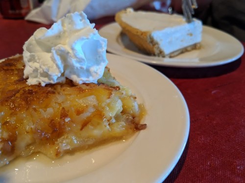 A slice of pineapple dessert topped with whipped cream, next to a slice of cream pie on a table.