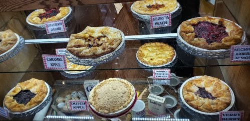 A display case filled with various pies, including peach, cherry, apple, and berry, each labeled with their names.