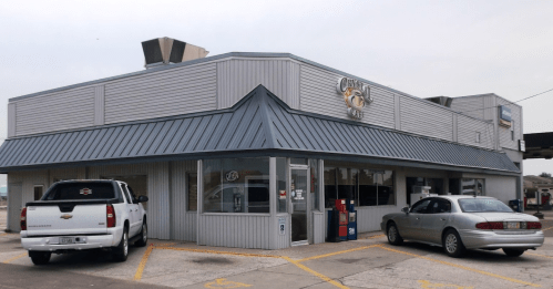 A gray building with a blue roof, featuring a restaurant sign, parked cars, and a clear sky in the background.