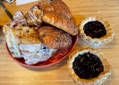 A plate of assorted pastries, including croissants and scones, alongside two small pies filled with dark berry jam.