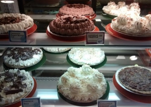 A display case filled with various cakes, including chocolate, coconut cream, and French silk, all topped with whipped cream.