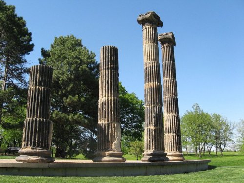Four weathered stone columns stand in a grassy area, surrounded by trees under a clear blue sky.