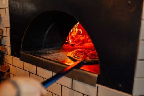 A pizza being placed into a wood-fired oven, with flames visible inside the oven.