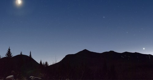 A serene night sky filled with stars, featuring a crescent moon and two bright planets above dark mountain silhouettes.