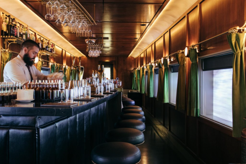 A bartender prepares drinks at a stylish bar with wooden interiors and green curtains, featuring a row of stools.