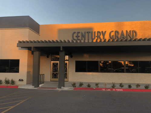 Exterior of a building with the sign "CENTURY GRAND," featuring a modern design and landscaped entrance.