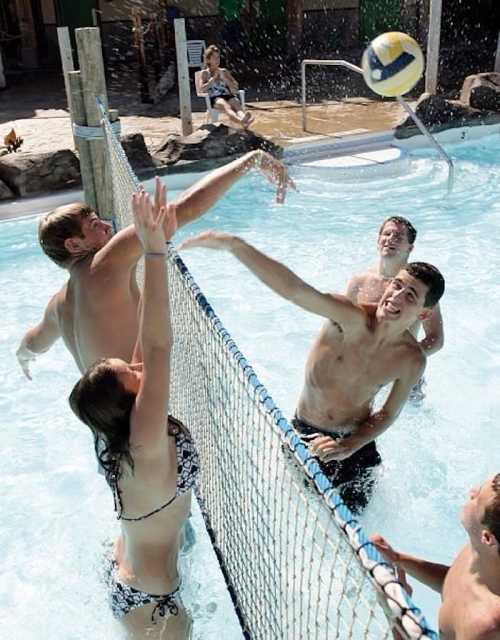 A group of young people playing water volleyball in a pool, with one player jumping to hit the ball over the net.