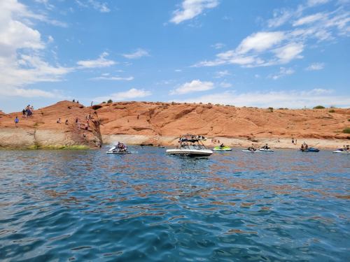 A scenic view of a rocky shoreline with people on boats and jet skis enjoying the water under a blue sky.