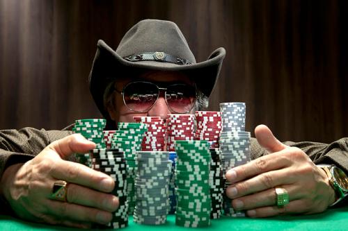 A man in a cowboy hat and sunglasses sits at a poker table, surrounded by stacks of colorful poker chips.